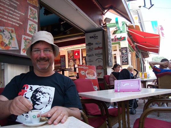 Man in hat drinking coffee at a café table, red chairs and awnings
