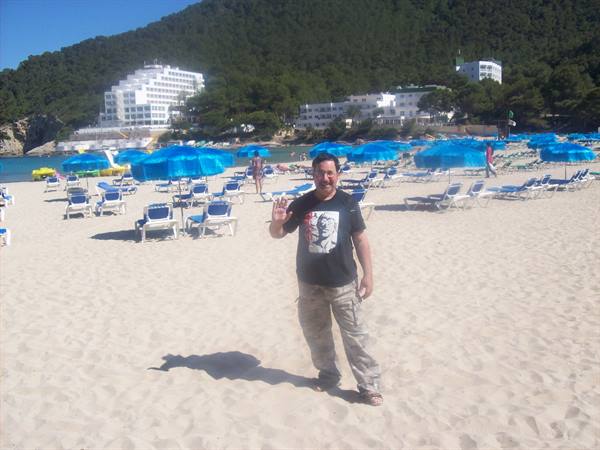 Man in black t-shirt waves on sandy beach with blue umbrellas and white hotels