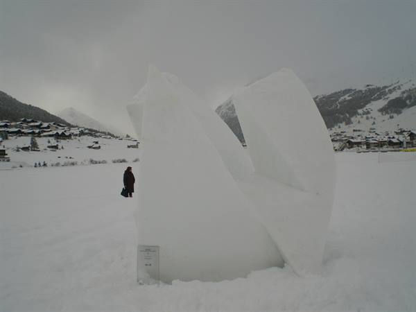 Snow sculpture on a snowy field, person nearby, village and mountains in the mist