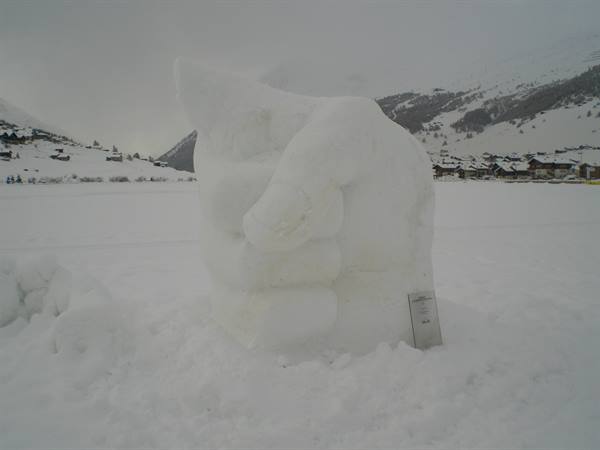 Snow sculpture shaped like a fist on a snowy plain, village and mountains under grey sky