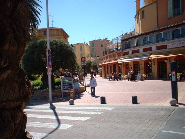 Woman with dog crossing paved square with cafés under orange awnings, colorful buildings