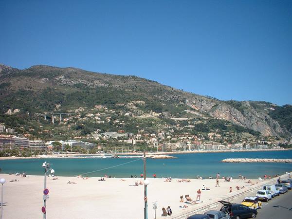 Beaulieu-sur-Mer beach: sunbathers on sand, turquoise sea, green mountains under blue sky