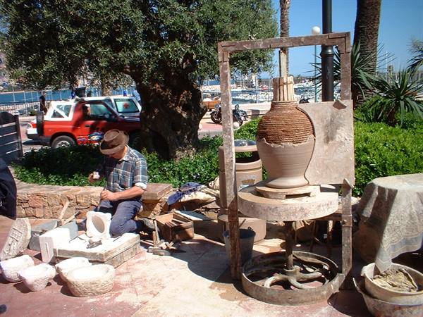 Potter shaping clay vessel on wheel near a marina with boats