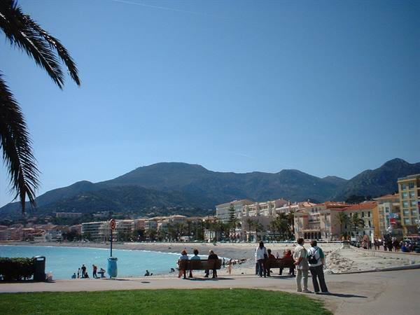 Tourists sitting and standing on sandy beach with colorful buildings and mountains