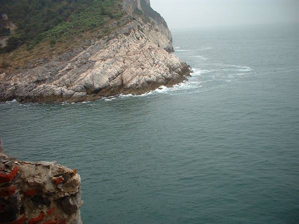 Rocky cliff drops into green sea, brick wall in foreground, hazy sky