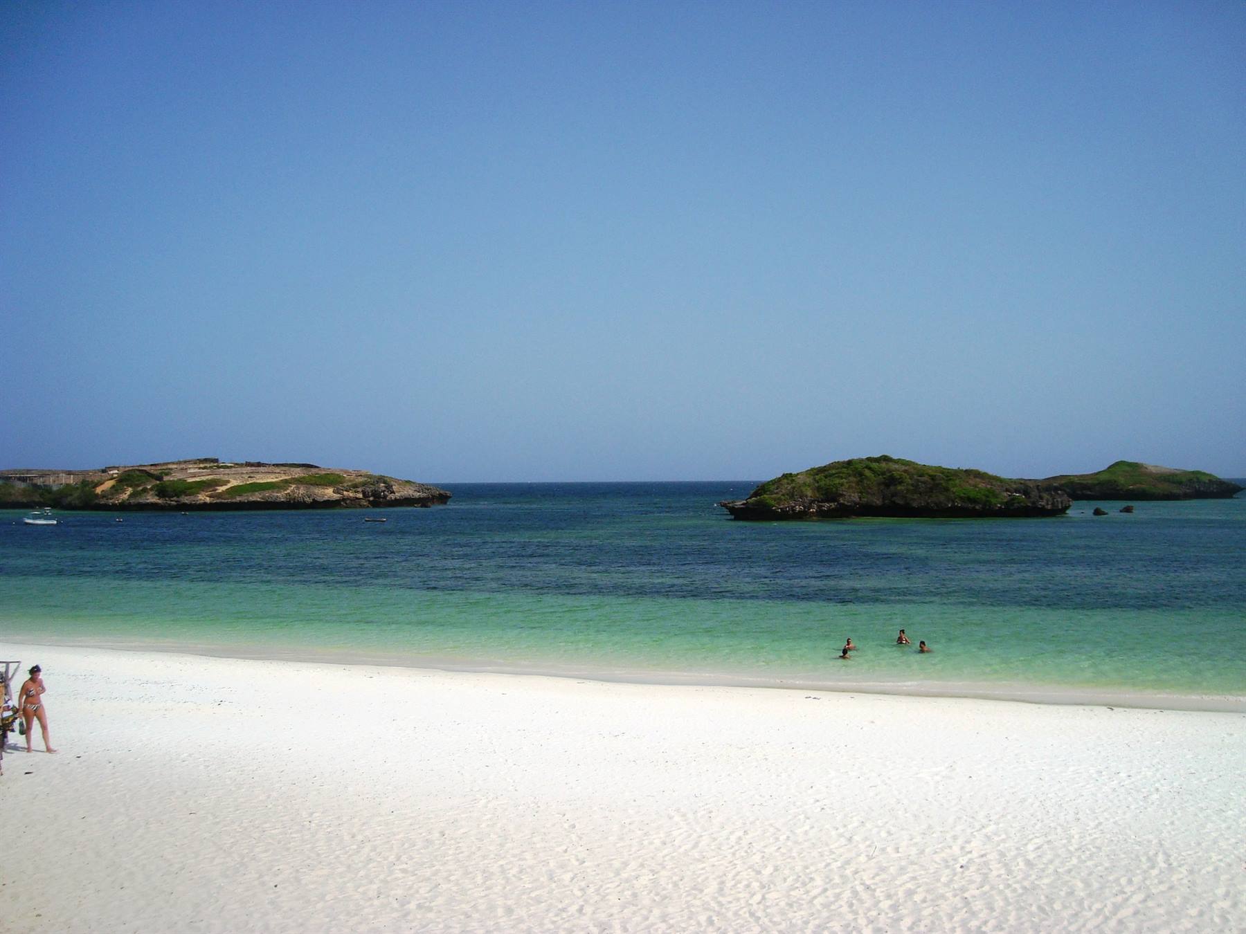 White sand beach, swimmers in turquoise water, green islets in the background
