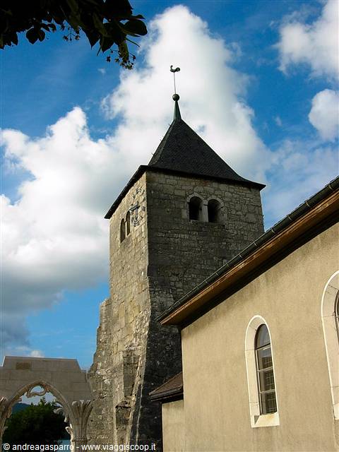 Stone bell tower with dark spire beside a church wall, white clouds above
