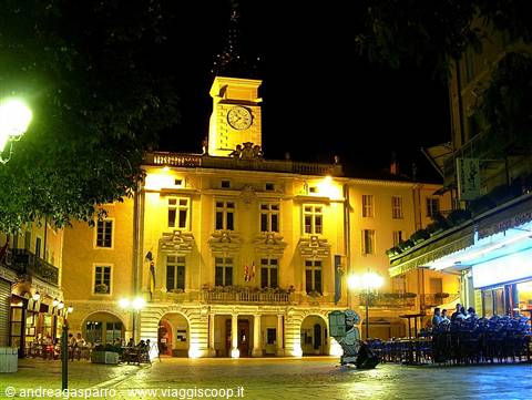 Night-lit town hall with clock tower, crowded café terraces on the square