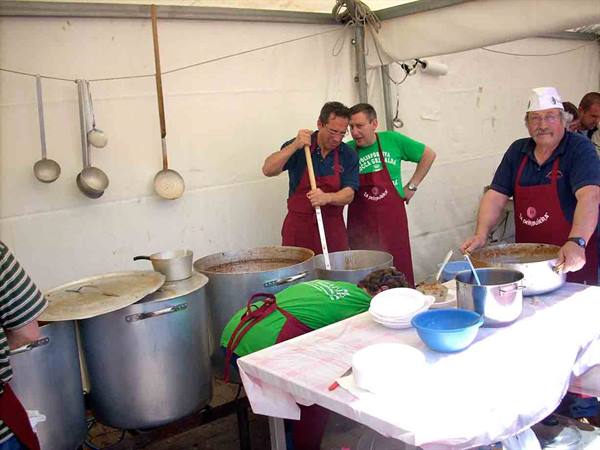 Cooks in red aprons stirring large metal pots under a white tent