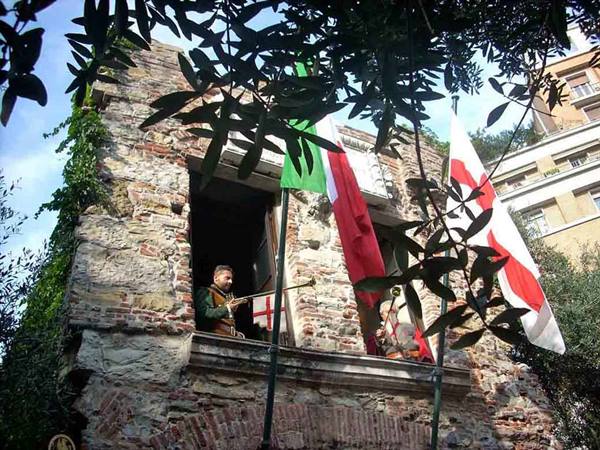 Trumpeter in costume on medieval stone balcony with Italian and Genoese flags