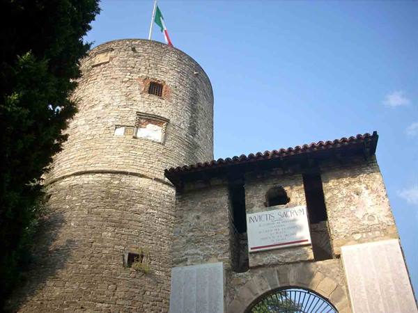 Medieval stone tower with Italian flag beside a stone gate with marble plaque