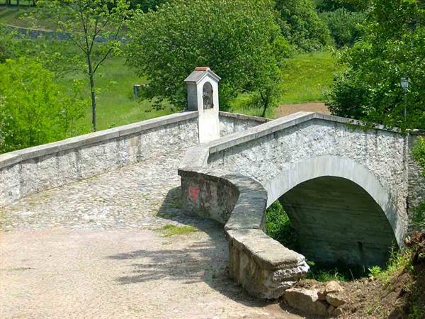 Single-arch stone bridge with small votive shrine, green meadows and trees
