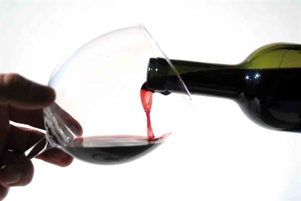 Red wine poured from green bottle into tilted glass on white background