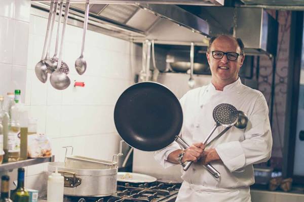 Smiling chef in white jacket holding pan and ladles in steel kitchen