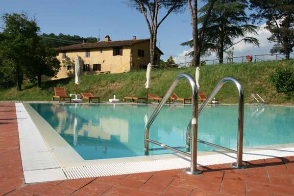 Steel-ladder pool in front of yellow farmhouse with umbrellas, loungers and trees