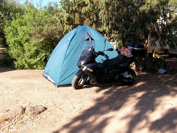 Blue tent and black scooter parked on sand, campsite among green trees