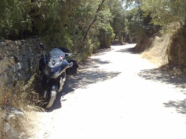 Black scooter parked beside a stone wall on a tree-lined dirt road