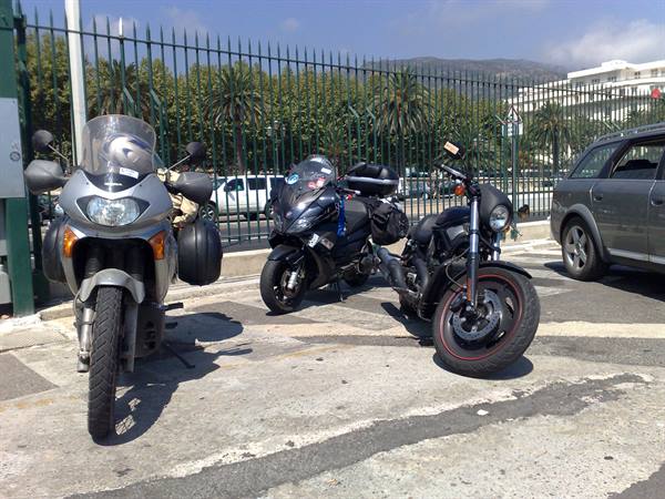 Three motorcycles parked beside a green fence with palm trees and mountains