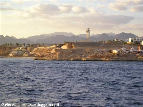 White lighthouse on rocky coast with desert mountains and blue sea