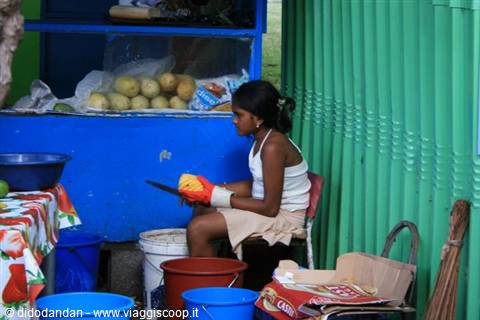 Woman seated beside blue fruit stall, colorful buckets and green corrugated wall