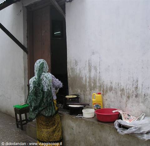 Woman in green headscarf cooking outdoors, pans and red basin on a wall