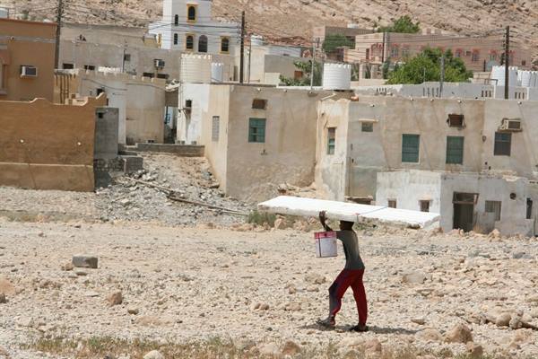 Man in red pants carries slabs on his head past whitewashed houses in arid village