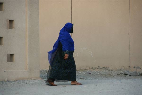 Woman in blue veil and black dress walking along ochre mud wall