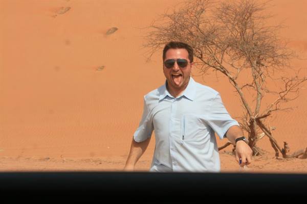 Man in sunglasses grimacing in front of a red dune and a dry tree