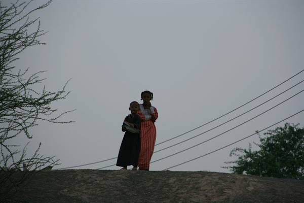 Two children embracing on a sandy hill, power lines and trees around