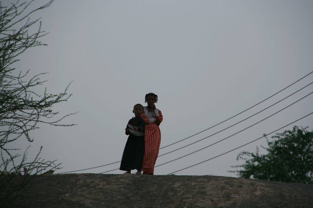 Two children embracing on a sandy hill, power lines and trees around