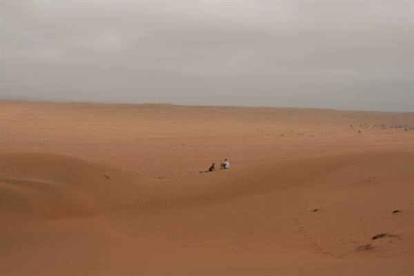Two people sitting on an orange sand dune under a grey sky