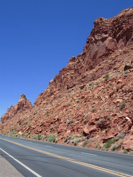 Empty highway beneath red sandstone cliffs, green shrubs, deep blue sky