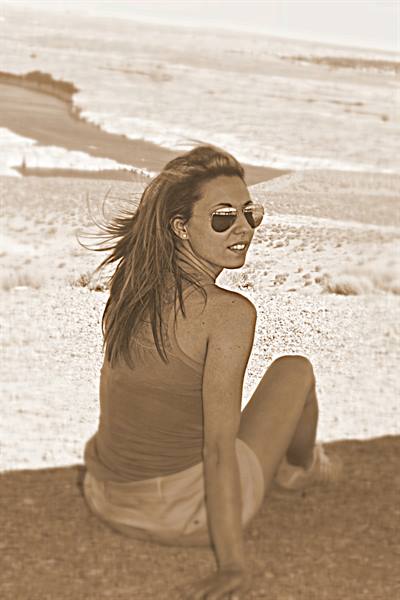 Woman in sunglasses sitting on the beach, hair blowing in the wind, sepia tone