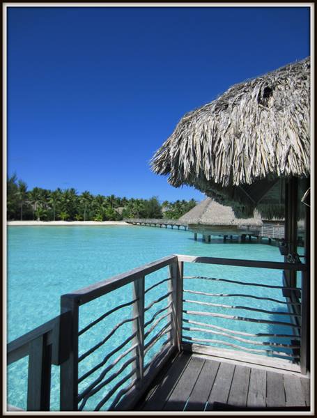 Wooden deck with thatched roof over turquoise lagoon, overwater bungalows nearby