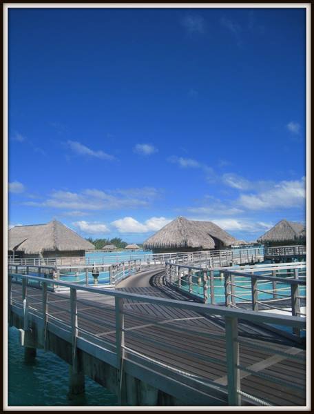 Wooden walkway leading to overwater thatched bungalows above turquoise lagoon, blue sky
