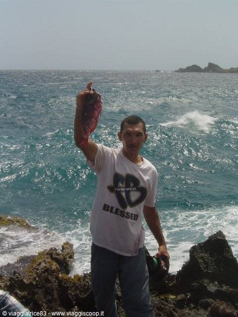 Fisherman holds up a red fish on black rocks, waves behind