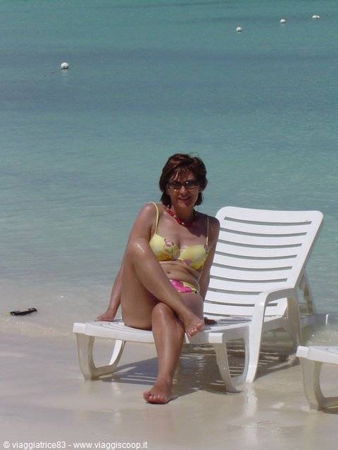 Woman in yellow bikini sits on white plastic lounger by turquoise shallow water