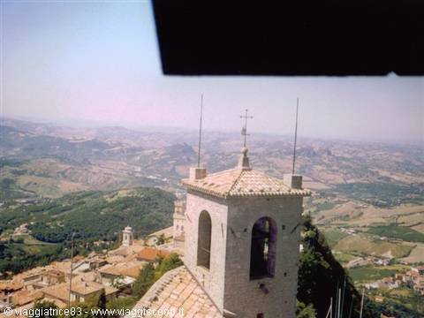 Stone bell tower with bell above rooftops, green rolling hills below, San Marino