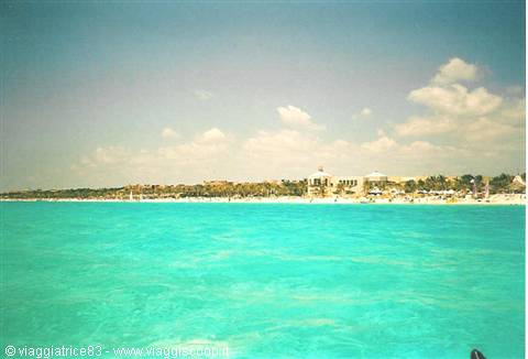 Turquoise sea in front of the beach with palm trees and hotels under white clouds