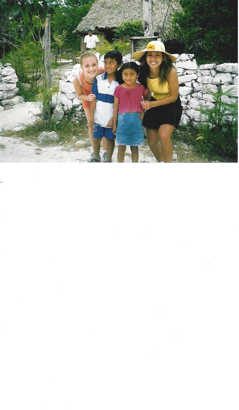 Four people smiling by a white stone wall and a thatched-roof hut