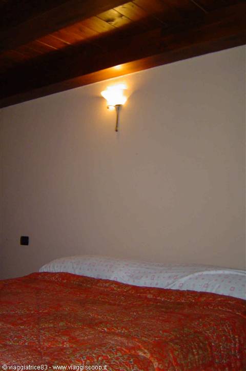 Room with orange bedspread, light wall and lit wall lamp under wooden beams