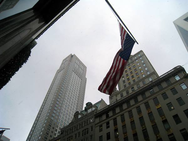 An American flag waves between Manhattan skyscrapers under a gray sky