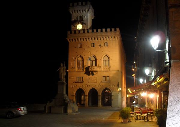 San Marino's illuminated Public Palace at night with clock, lanterns and café tables