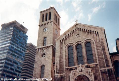 Stone church with clock tower rising between glass skyscrapers under clouds