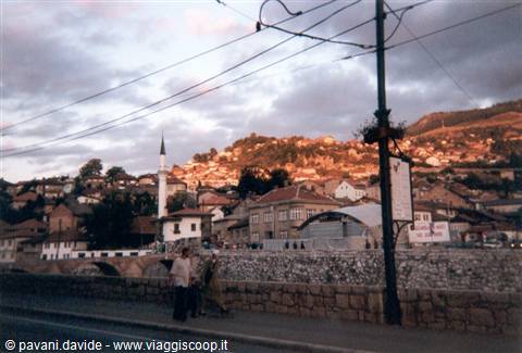 Sarajevo: minaret and hillside houses at dusk, pedestrians by the river
