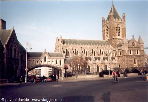 Christ Church Cathedral in Dublin, grey stone, covered bridge and cyclists under sunny sky