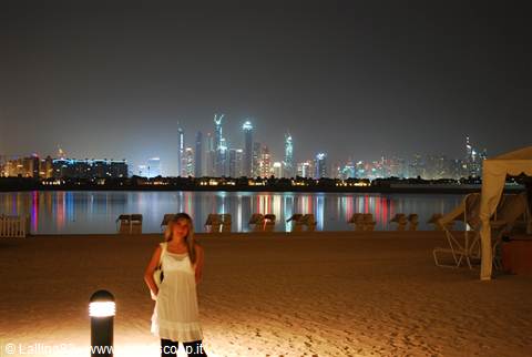 Woman in white dress on night beach, illuminated Dubai skyscrapers reflected on water