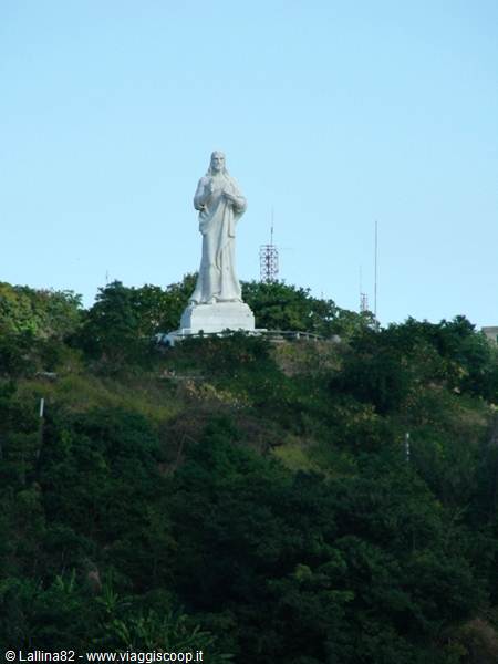 White Christ statue on green hill, antennas among trees, clear sky