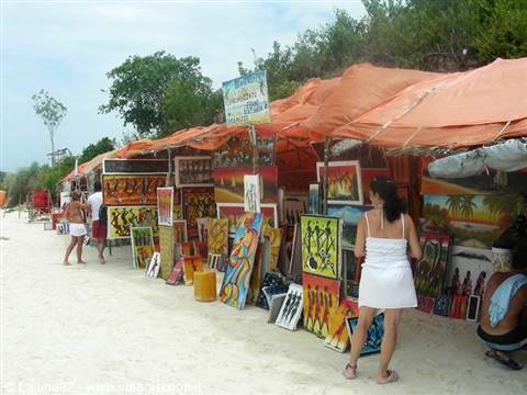 Woman in white dress browsing paintings for sale under orange tarps on the beach