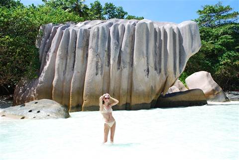 Woman in bikini wading in shallow water under huge granite boulder, La Digue